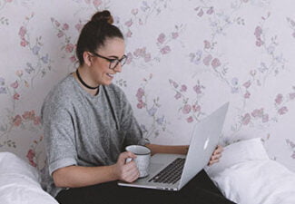 Kvinna som sitter med laptop och kaffekopp i sin säng