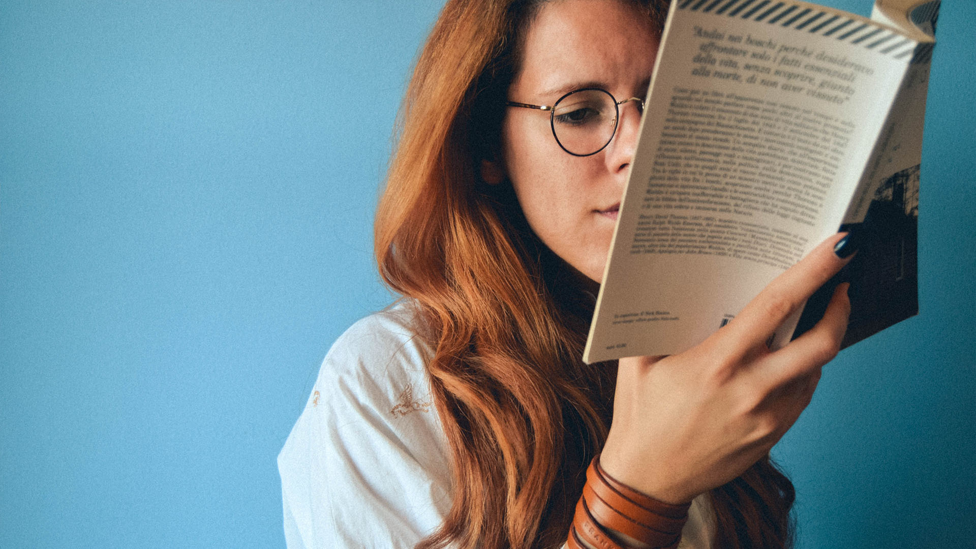Rödhårig kvinna med rundade glasögon som håller i en bok som täcker halva ansiktet