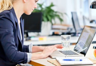 Kvinna i blå kavaj och uppsatt hår som sitter vid laptop och arbetar
