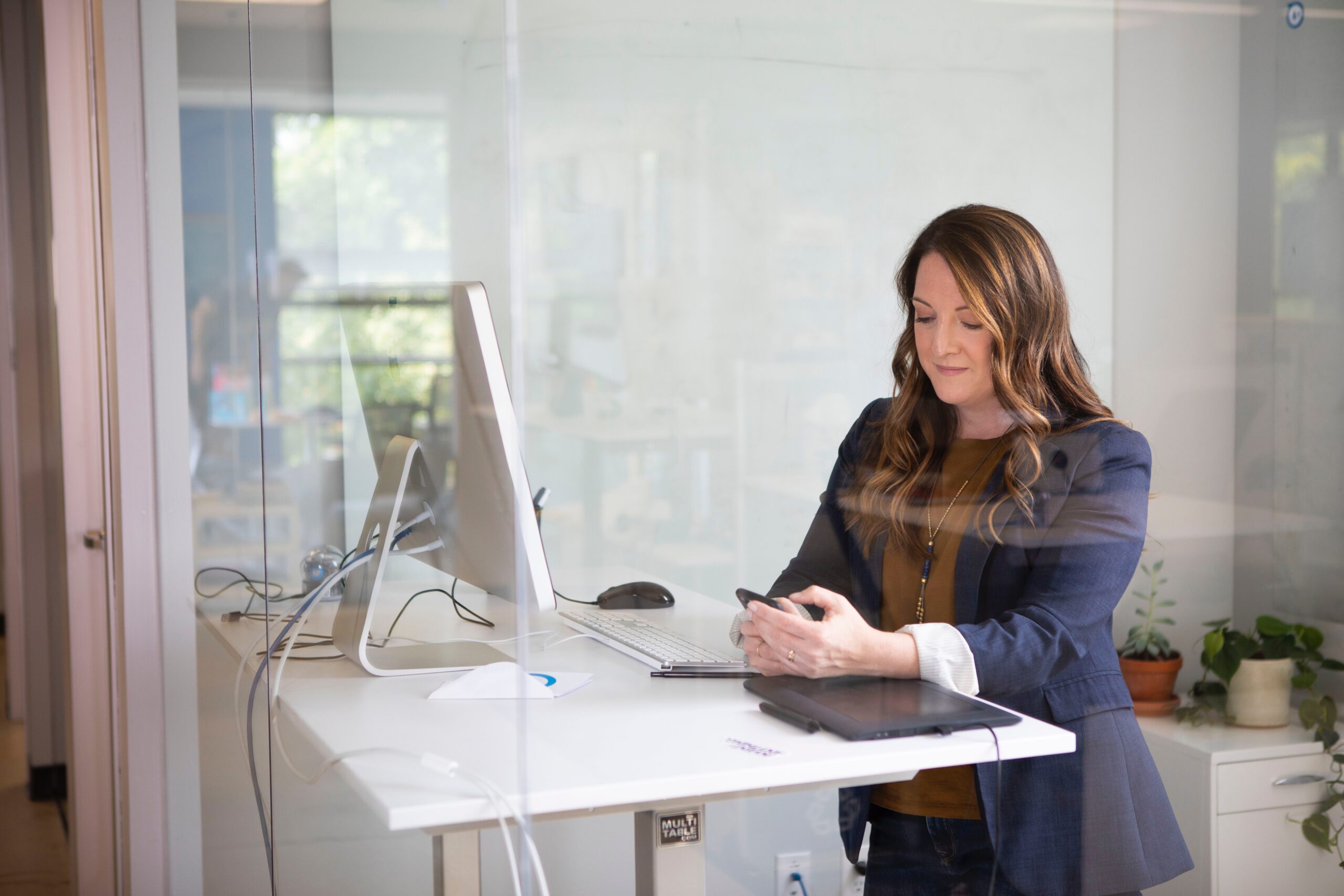 Kvinna i blå kavaj som arbetar vid laptop och mobil, stående vid sitt skrivbord på kontoret med glasväggar
