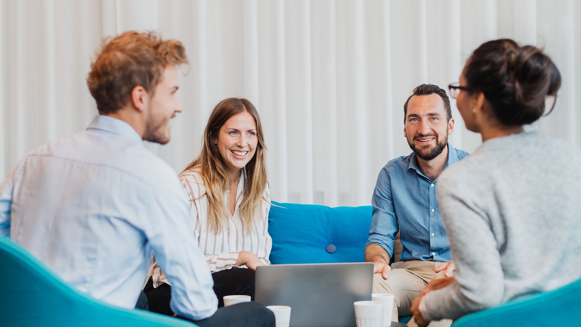 Fyra personer som sitter i en soffgrupp på ett kontor och pratar över en kopp kaffe.