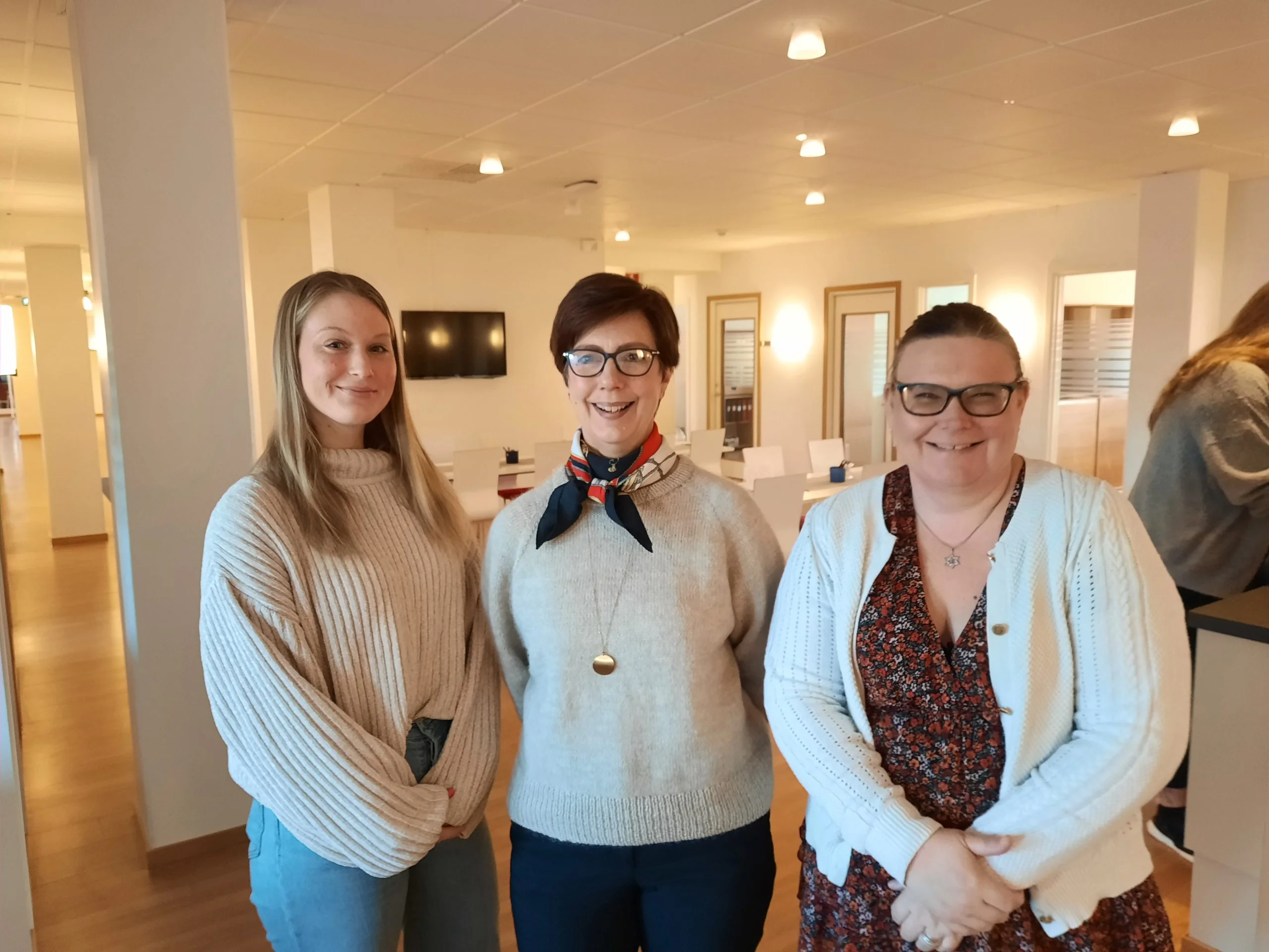 Tre personer står tillsammans i ett ljust och modernt rum. Den första personen till vänster har långt blont hår och bär en stickad tröja. Personen i mitten har kort hår, glasögon och en scarf runt halsen. Personen till höger har glasögon och en vit kofta över en blommig klänning. I bakgrunden syns möbler och en stor skärm på väggen.
