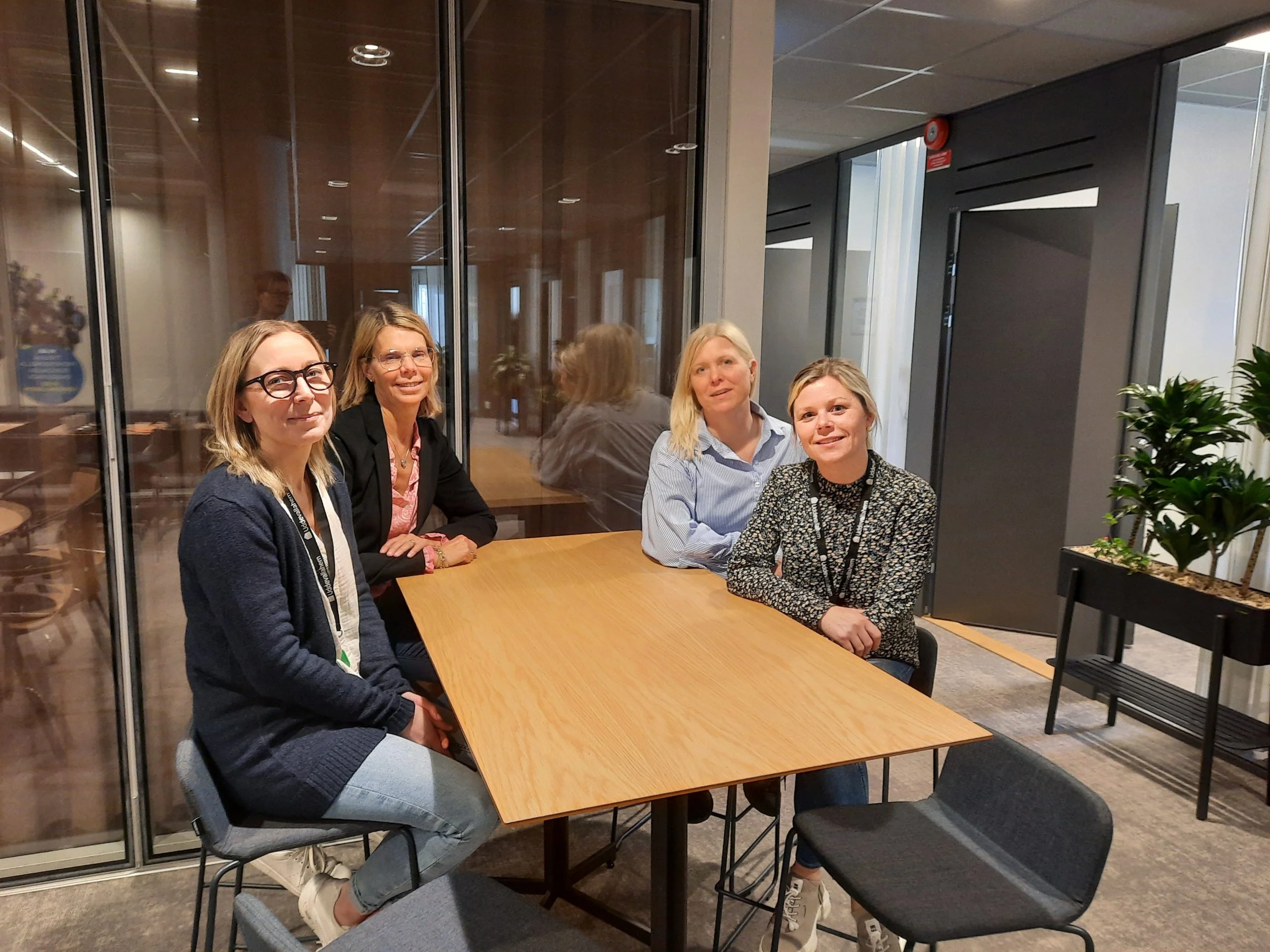 Fyra personer sitter runt ett rektangulärt träbord i ett modernt mötesrum. Till vänster syns en kvinna med glasögon, mörk kofta och jeans som ler mot kameran. Bredvid henne sitter en kvinna i svart kavaj och rosa skjorta, följd av en kvinna i ljusblå skjorta med blont hår. Längst till höger sitter en kvinna i mönstrad tröja och jeans. I bakgrunden finns glasväggar och gröna växter som skapar en ljus och professionell atmosfär.