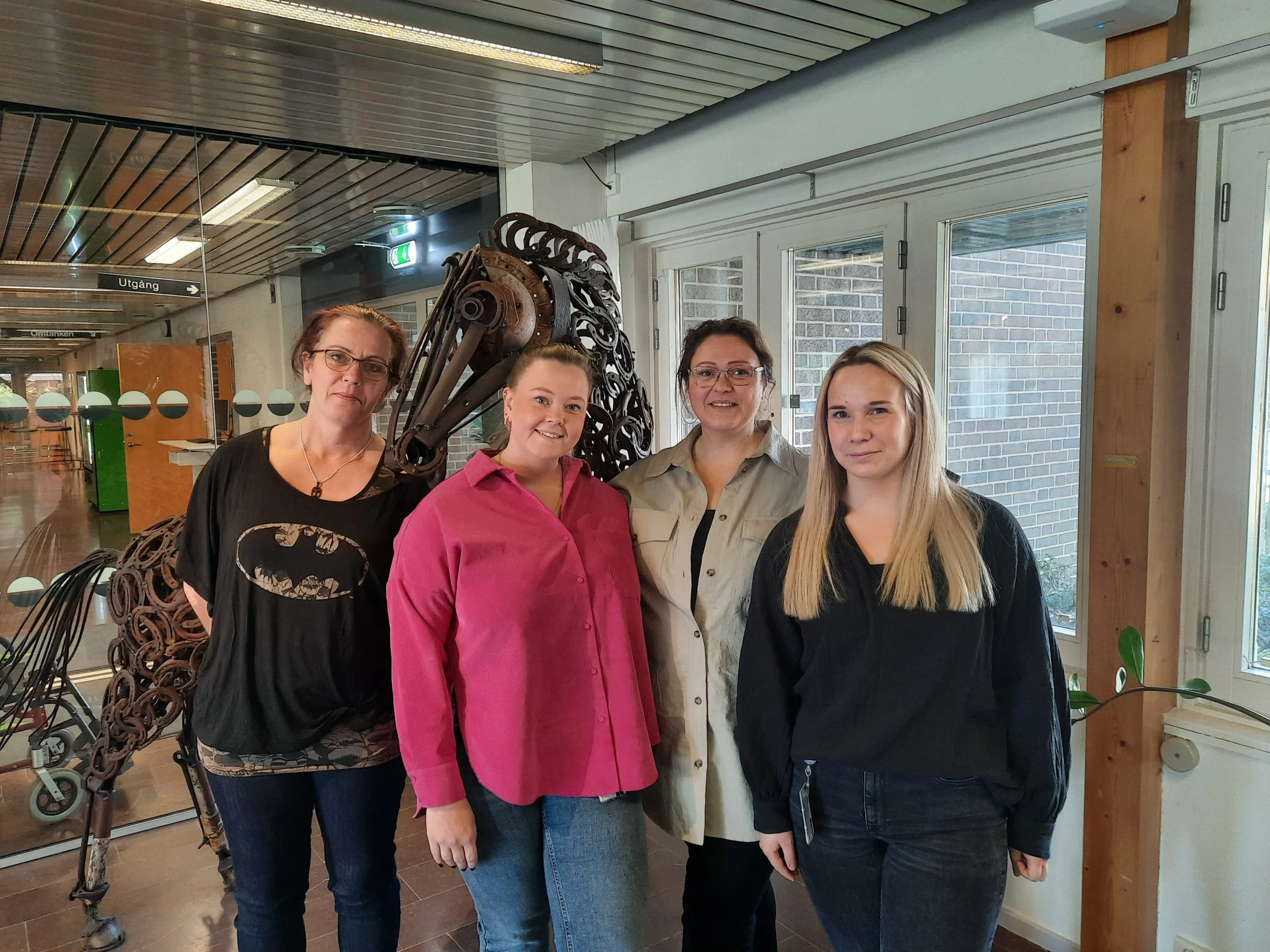 Fyra kvinnor står bredvid varandra i en ljus inomhusmiljö. Till vänster står en kvinna med glasögon och en svart Batman-tröja, följt av en kvinna i en rosa skjorta. Bredvid henne står en kvinna i beige skjorta och glasögon, och längst till höger en kvinna med blont hår och svart blus. Bakom dem syns glasfönster, en skulptur av en häst i metall samt trädetaljer som ger rummet en varm och kreativ atmosfär.