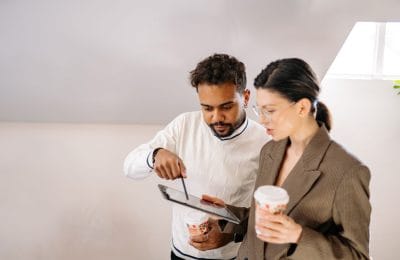 Två kollegor diskuterar och pekar på en surfplatta medan de håller kaffemuggar.